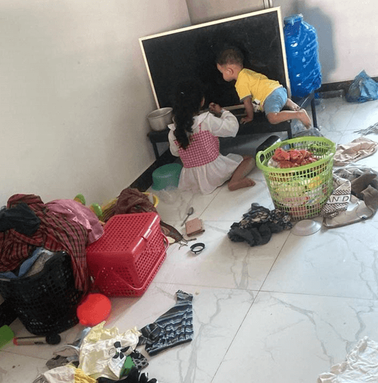 An 8-year-old girl and a 3-year-old boy drawing on a small blackboard in a messy room with laundry scattered around.