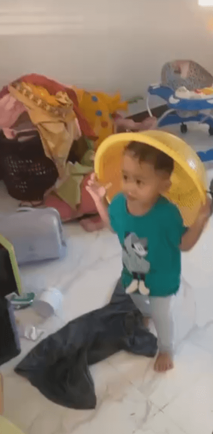 A 3-year-old boy wearing a yellow plastic colander on his head like a helmet in a messy room.