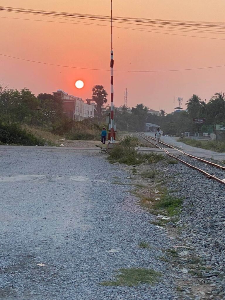 A bright orange sun setting over a gravel railway crossing and train tracks, with a few locals walking nearby in the late afternoon.