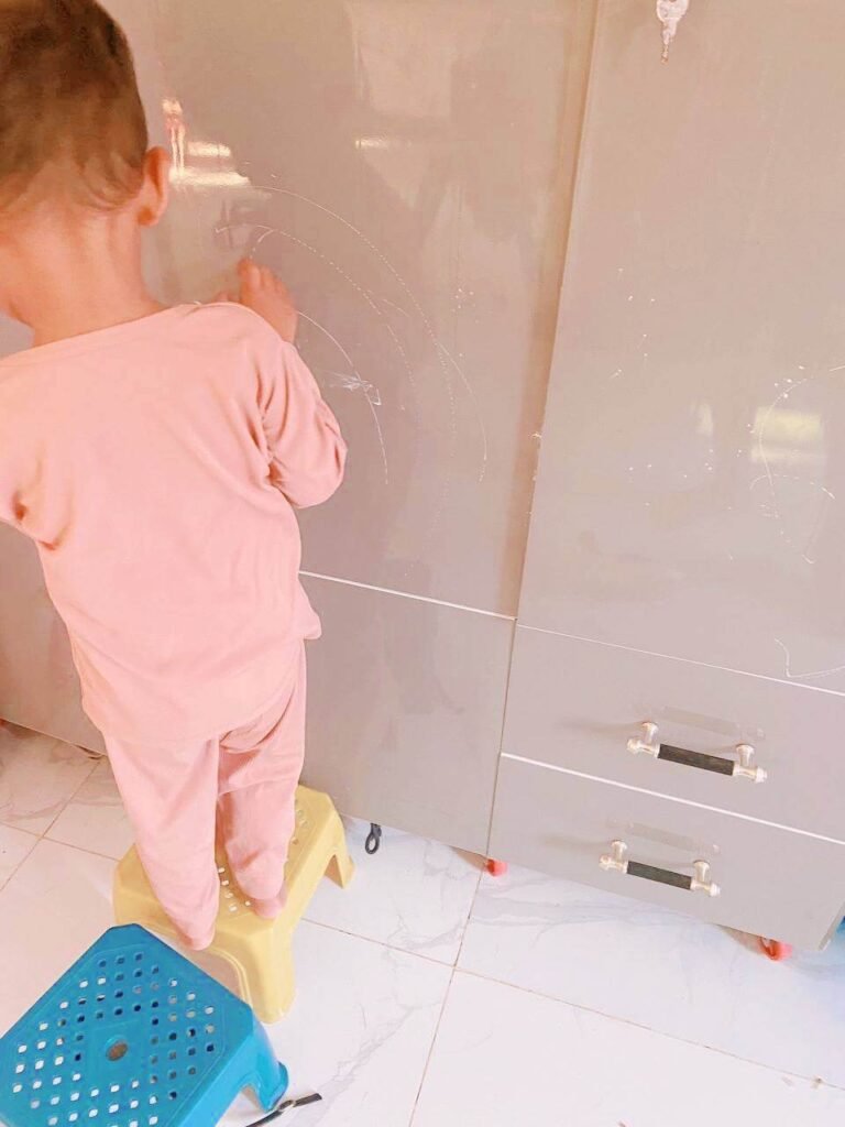 A young boy balancing on a yellow plastic stool next to a blue stool, touching the surface of a grey cabinet.