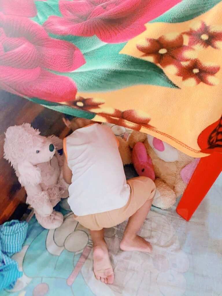 A toddler crawling on his hands and knees under a colorful floral blanket next to two large stuffed animals.