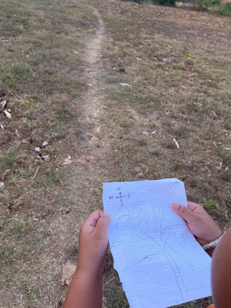 A child's hands holding a hand-drawn paper map with a compass rose while standing on a dry dirt path.