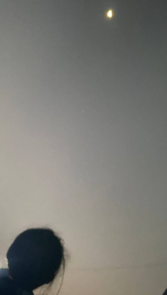 The silhouette of a child's head looking up at a glowing crescent moon in a hazy evening sky.