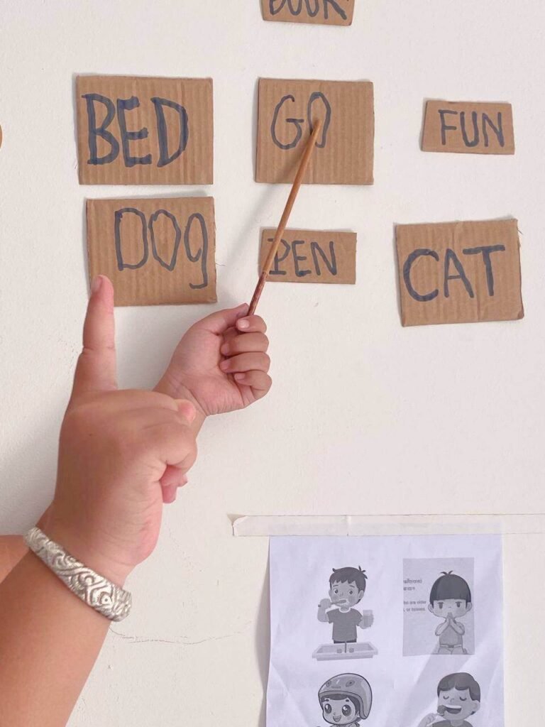 A child's hand using a pointer stick to select vocabulary words like bed, dog, and cat written on cardboard squares taped to a wall.