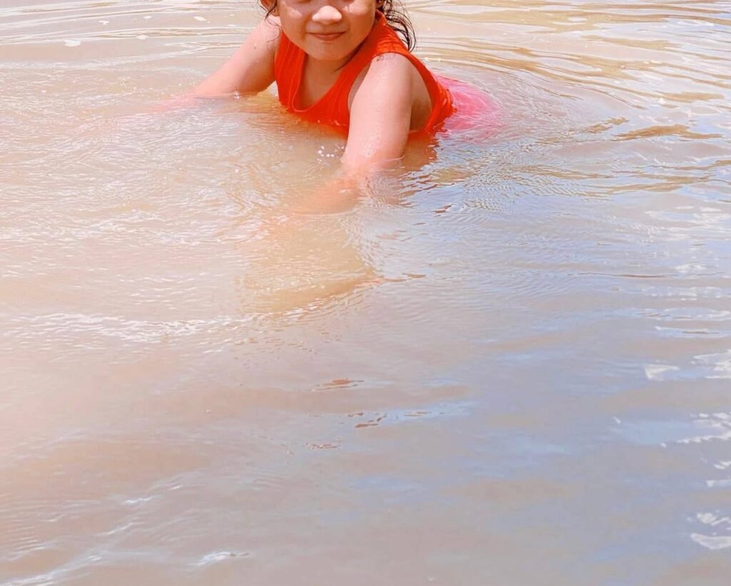 A young girl in an orange top smiling while playing in the shallow, murky water of a natural stream.