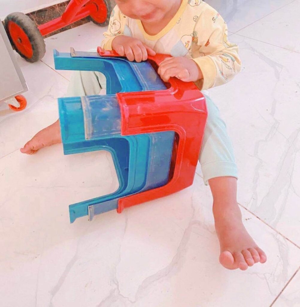 A toddler sitting on the floor working to pull apart a stack of red and blue plastic step stools.