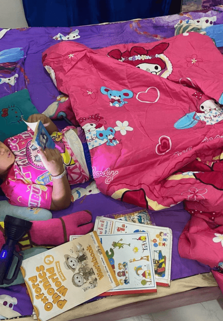 A child lying under a pink blanket in bed, reading a small book with a black flashlight resting nearby.