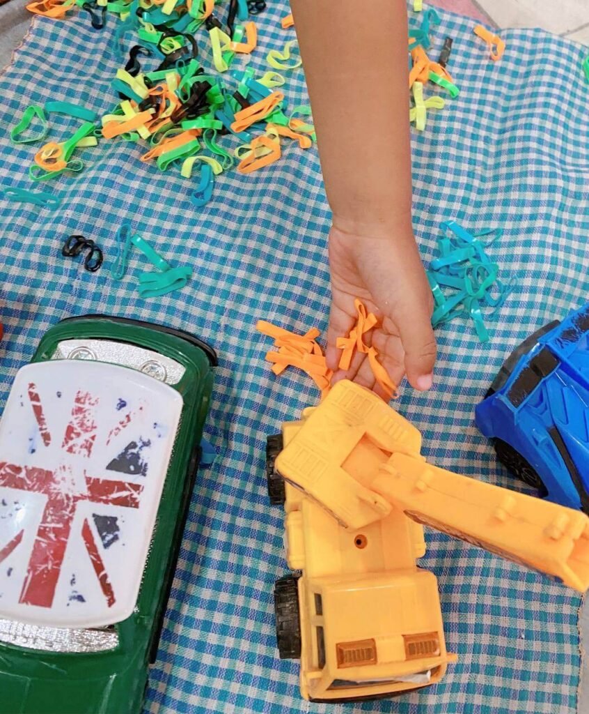 A child's hand dropping orange rubber hair ties onto a yellow toy crane, surrounded by green and blue toy cars and piles of colored bands on a checkered mat.