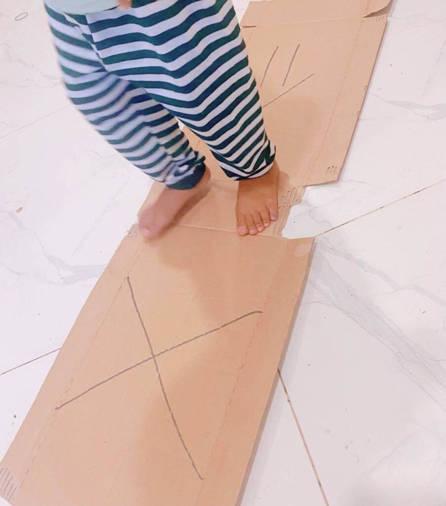 A toddler's bare feet stepping on flattened cardboard pieces that have a large X and an arrow drawn on them in marker.