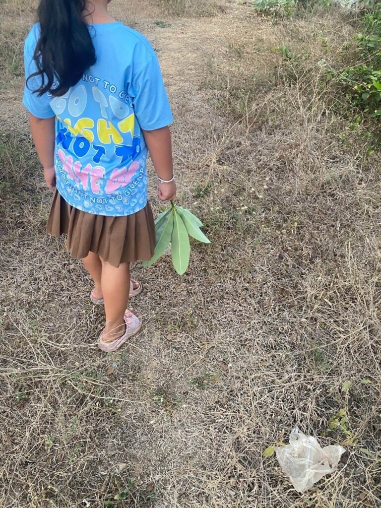 A young girl in a blue shirt walking away on a dry, overgrown dirt path while holding green leaves.
