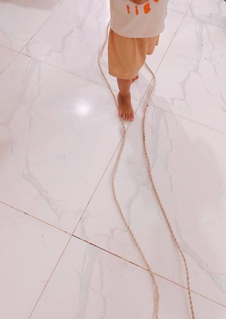 A toddler's bare feet carefully walking between two pieces of thick rope laid parallel on a tiled floor.