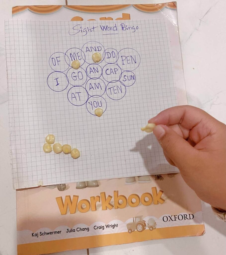 A child using dried corn kernels as markers on a homemade bingo board filled with short vocabulary words.