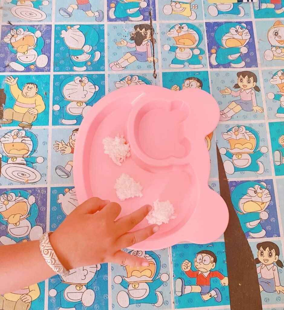 A child’s hand reaches for three small piles of cooked white rice on a pink plate, set against a blue Doraemon patterned tablecloth.