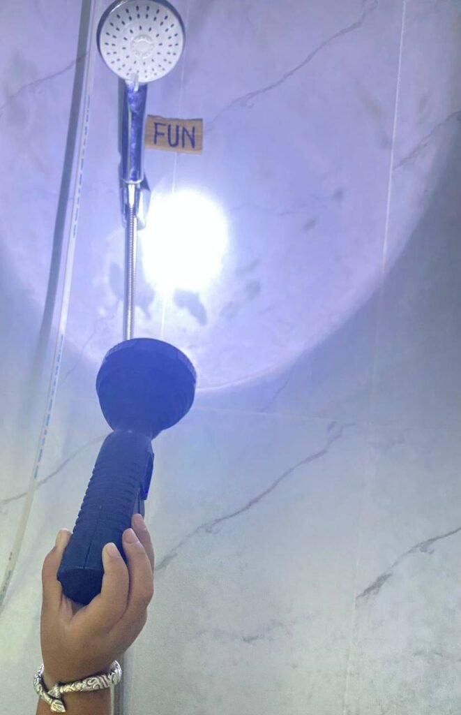 A child holding a blue flashlight and shining the beam onto the word FUN taped to a tiled bathroom wall near a showerhead.