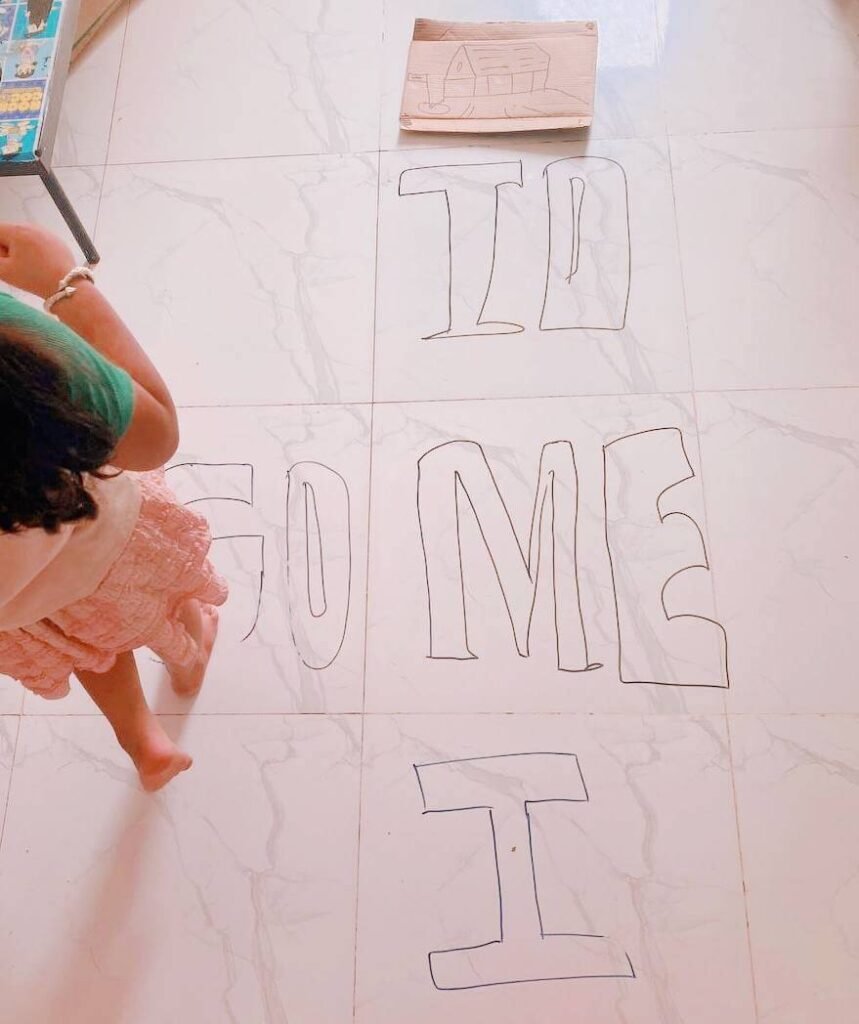 A young child's feet standing on a tiled floor where the words TO, ME, and I are drawn out to look like a hopscotch grid.