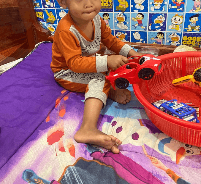 A toddler sitting on a bed, happily putting a red toy car into a red plastic basket during a cleanup game.