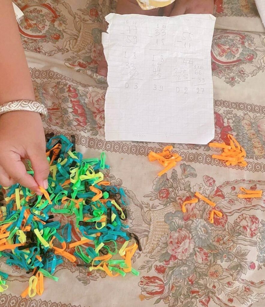 Overhead view of a child's hand selecting colorful rubber hair ties next to handwritten subtraction problems on a floral bedspread.