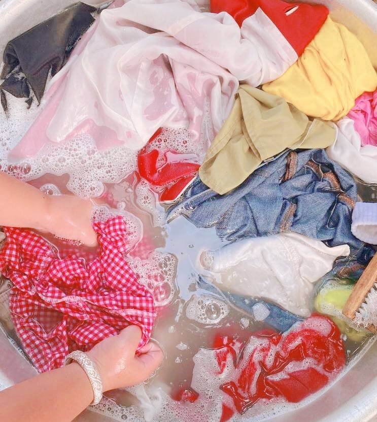 A child's hands washing small items of clothing in a large metal basin filled with soapy water.