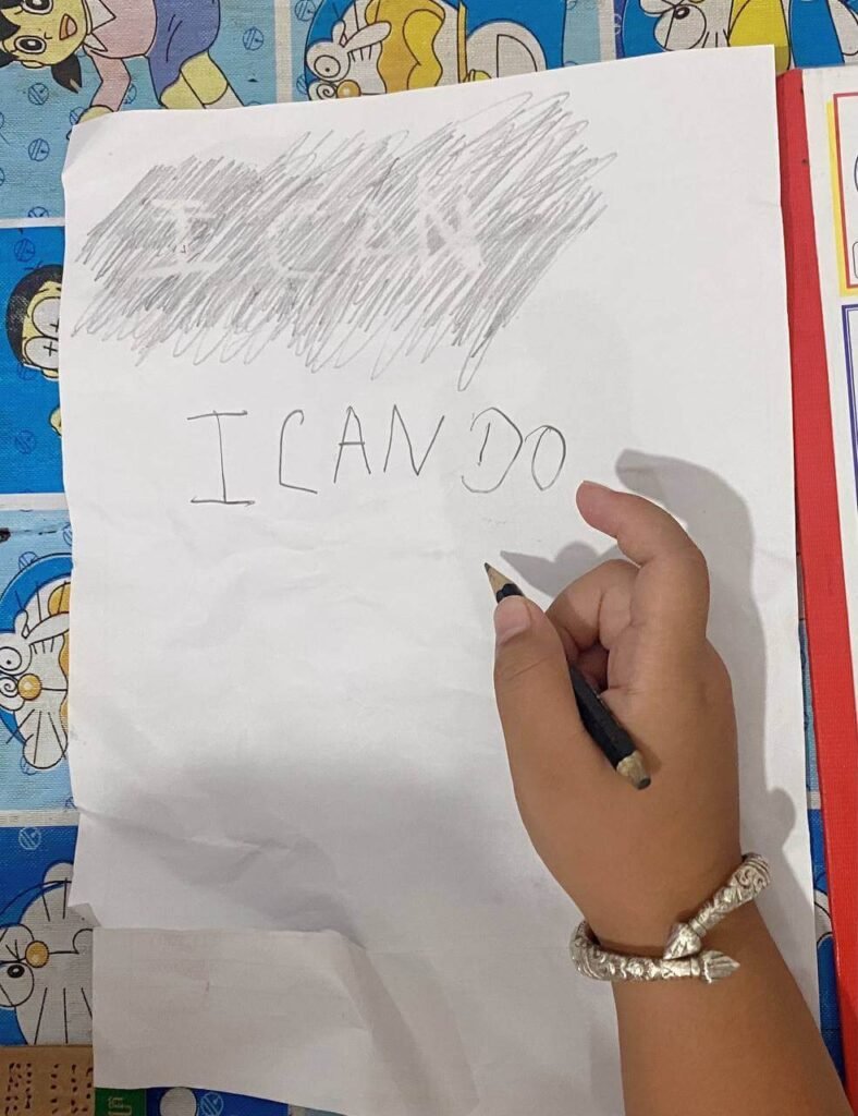 A child using a dark pencil to shade over white paper, revealing the hidden phrase "I CAN" and adding their own words below it.