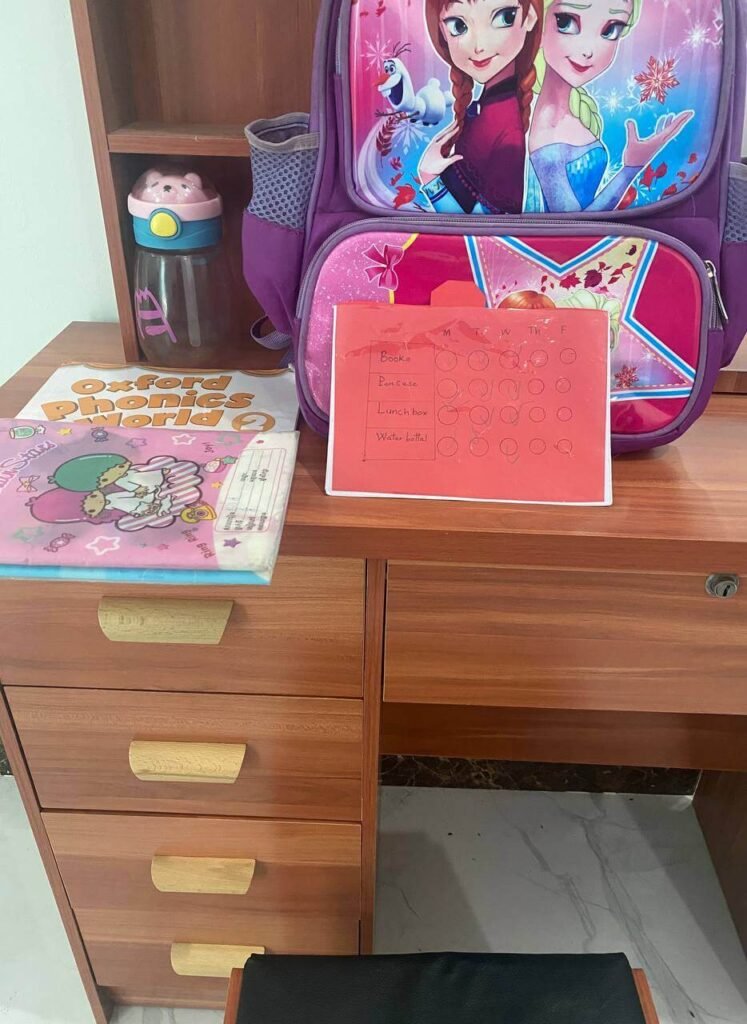 A purple school backpack sitting on a wooden desk next to a handmade red paper checklist for packing daily school supplies.