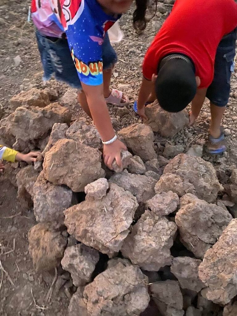 Two young children bending down to carefully stack large, dry clumps of earth into a pile on the dirt ground.