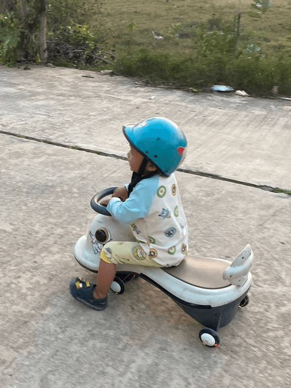 A young boy wearing a blue helmet, sitting on a small ride-on toy outdoors on a paved driveway.