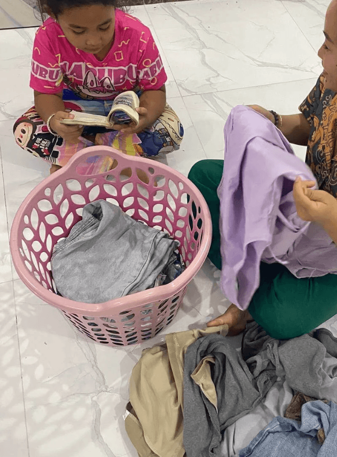 A mother folding clothes from a pink laundry basket on the floor while her daughter sits across from her reading a book.