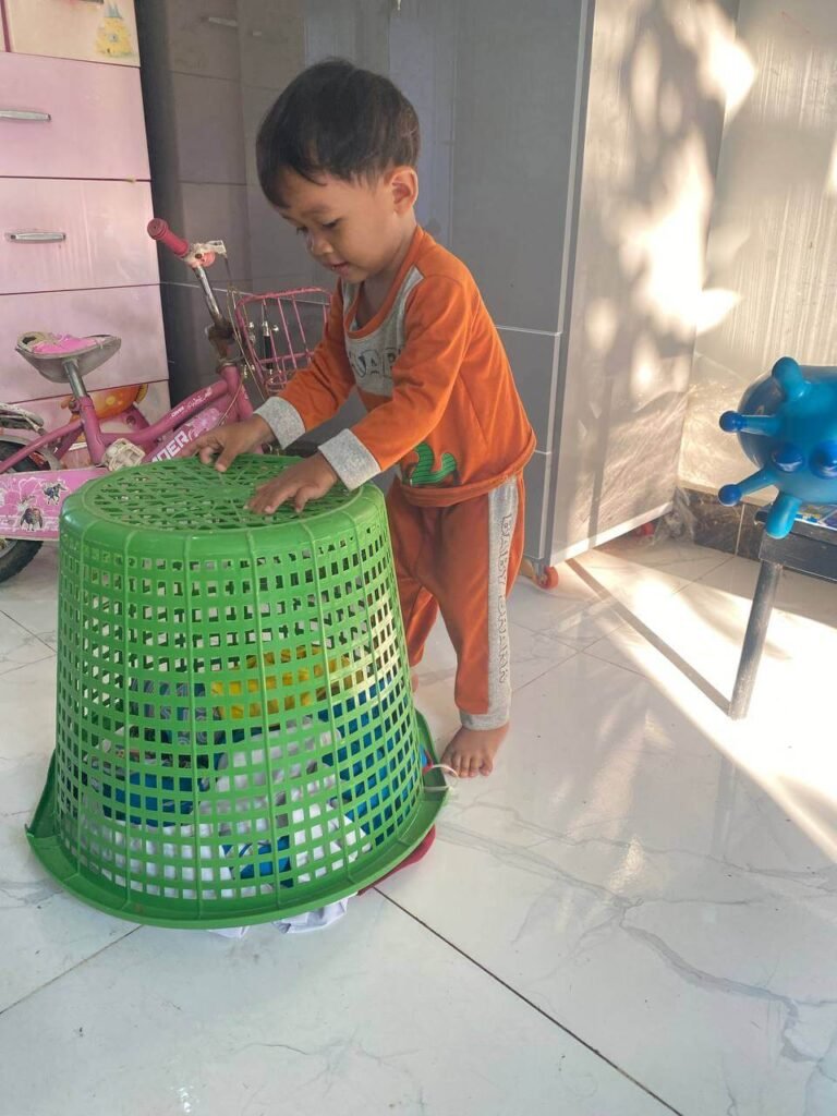 A young boy standing next to a large green laundry basket that has been turned upside down over clothes on a tiled floor.