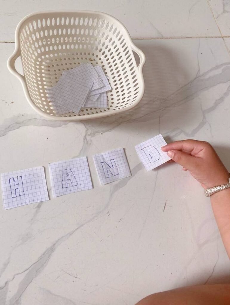A child placing small squares of grid paper with letters drawn on them side-by-side on the floor to spell the word HAND.