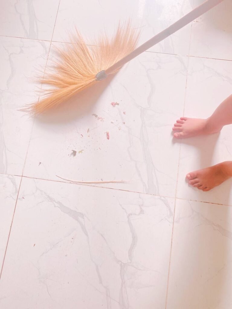 A child's bare feet next to a straw broom sweeping a small pile of dust on a white tiled floor.