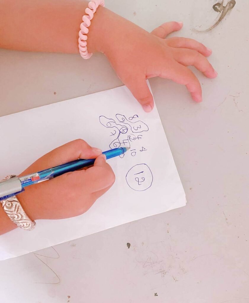 Close up of a child's hand holding a blue pen and circling numbers drawn on white paper as part of a math game challenge.