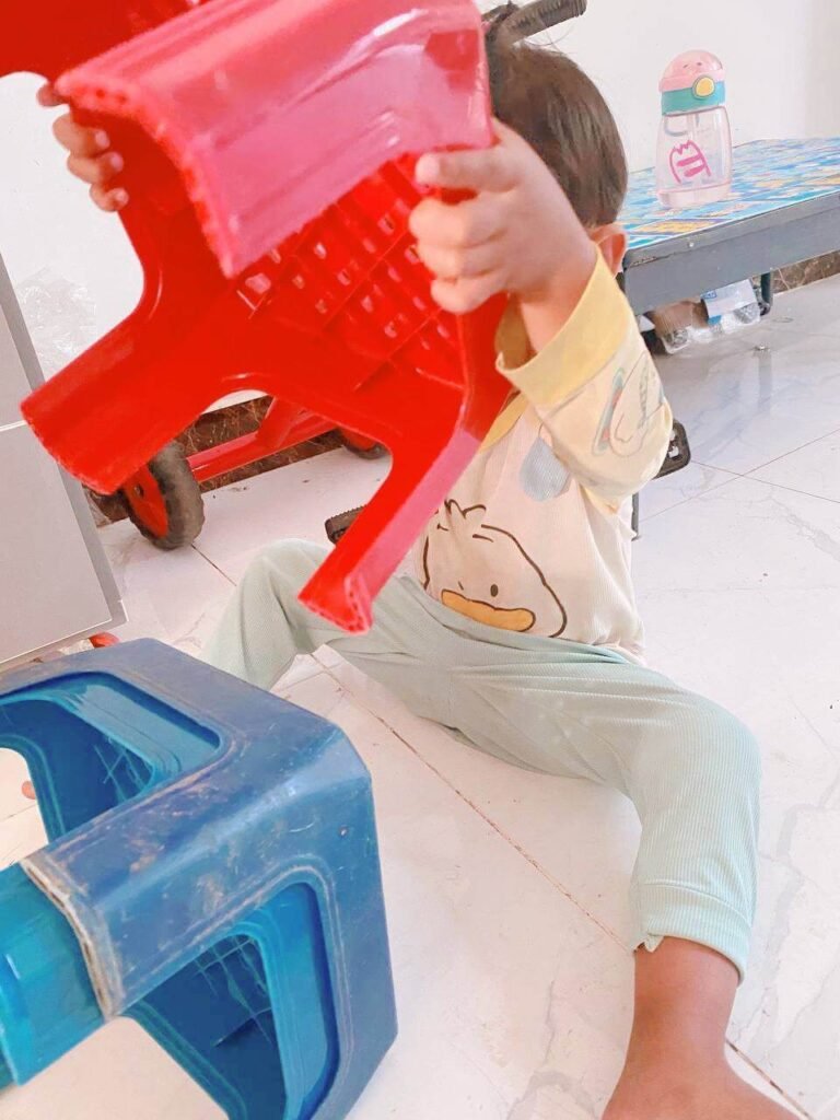A toddler sitting on a tiled floor looking frustrated while trying to pull apart red and blue plastic stools.