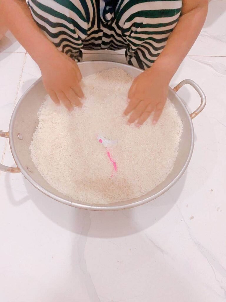 A child's hands digging through a large metal pan filled with uncooked white rice to find a small hidden toy.