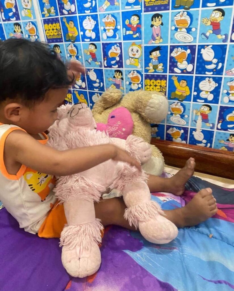 A toddler sitting on a colorful mat carefully holding a large pink stuffed poodle toy.