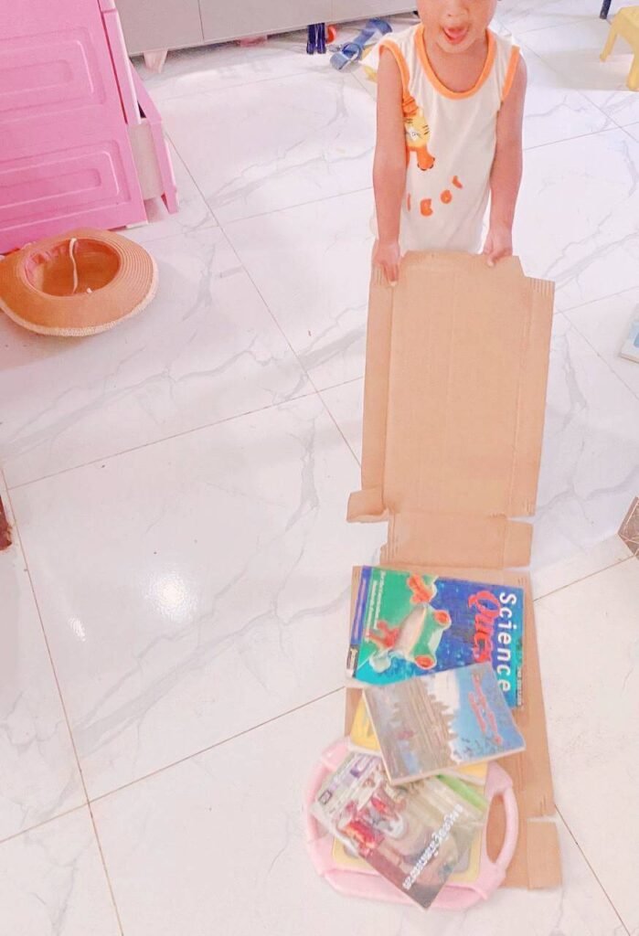 A young boy standing on a tiled floor pulling a piece of cardboard loaded with children's books.