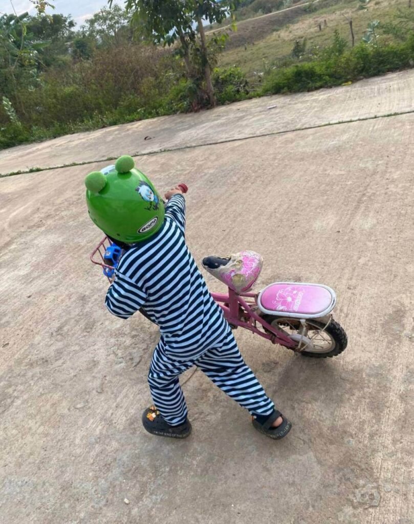 A young boy wearing a green frog helmet pushing a pink pedal bike along a concrete path.