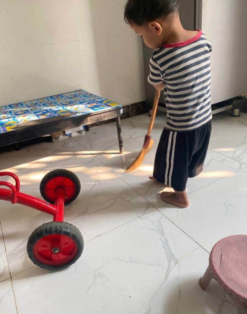 A young boy using a small broom to sweep the tiled floor next to a red toy tricycle.