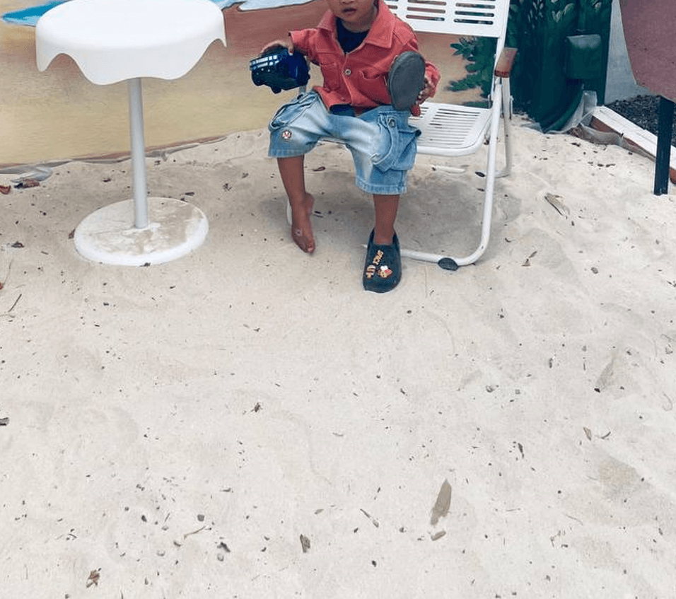 A toddler sitting on a chair in an outdoor sandbox area, holding a shoe and getting ready to transition away from playtime.