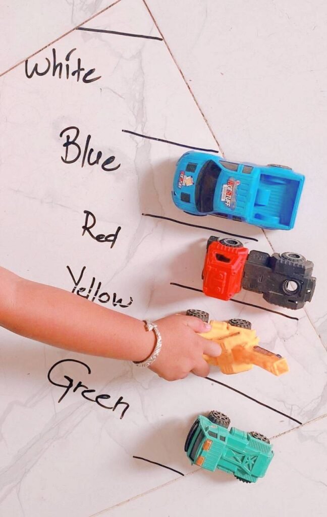 A child's hand driving small toy cars into a hand-drawn parking lot where each space is labeled with color words like blue, red, and yellow.