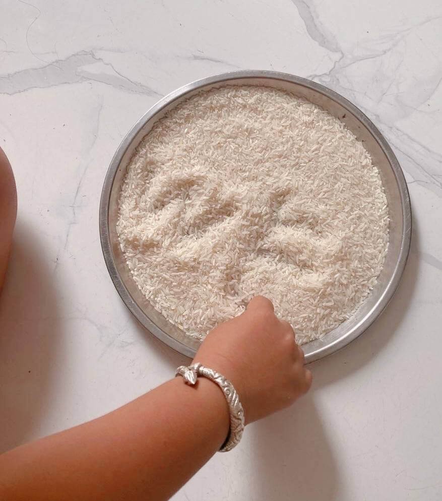 A child's hand tracing letters with their finger inside a round metal baking tray filled with uncooked white rice.