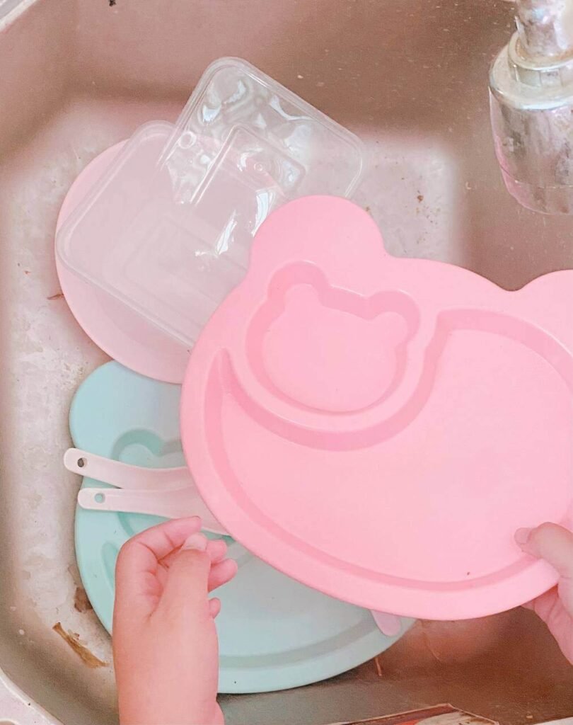 A child's hands washing and organizing a pink plastic bear-shaped plate and other lightweight plastic containers in a sink.