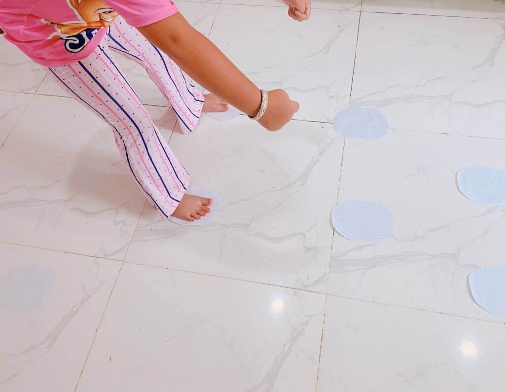 A child standing barefoot on a tile floor, stepping specifically onto white paper plates arranged as stepping stones.