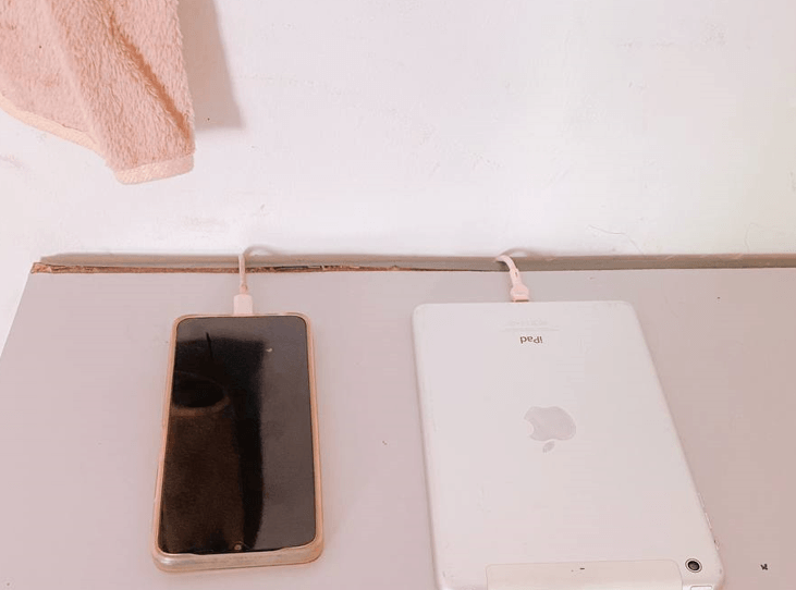 An iPhone with a gold case and an Apple iPad plugged into white charging cables side-by-side on a central charging desk.