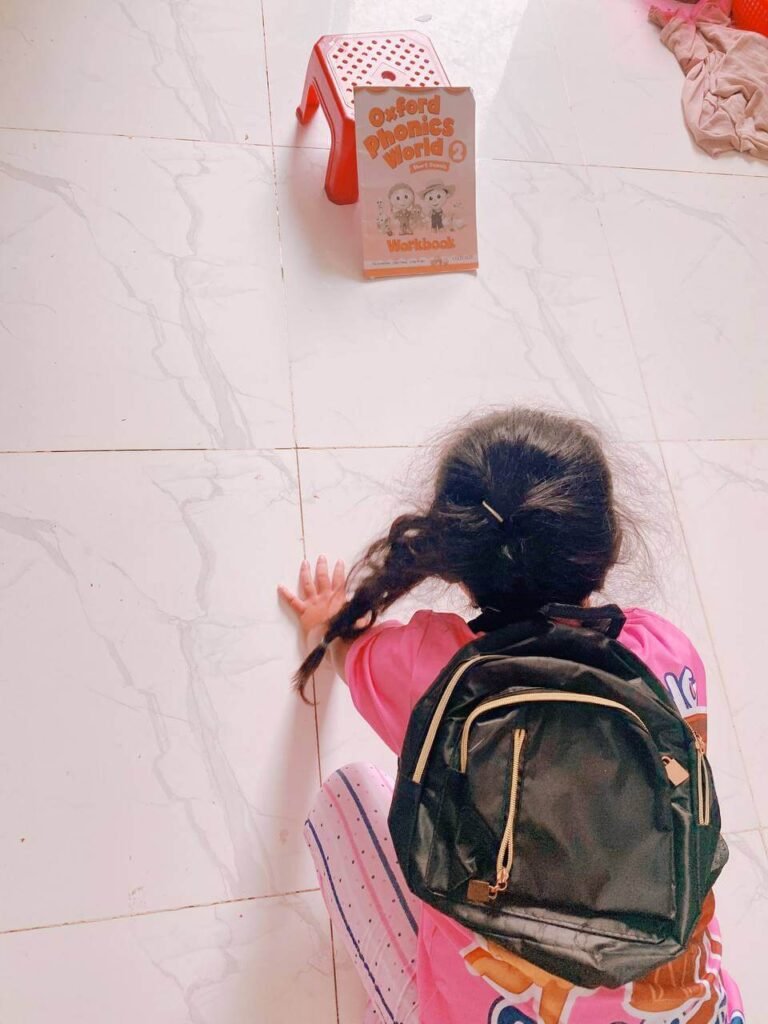 An older girl facing away from the camera, wearing a backpack, with her hand near a propped-up "Oxford Phonics World 2 Workbook" next to a red stool.