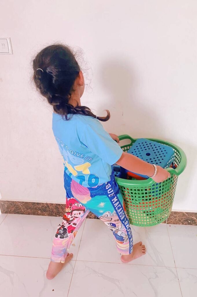 A child carrying a large green plastic laundry basket filled with a blue stool and toys across a tile floor.