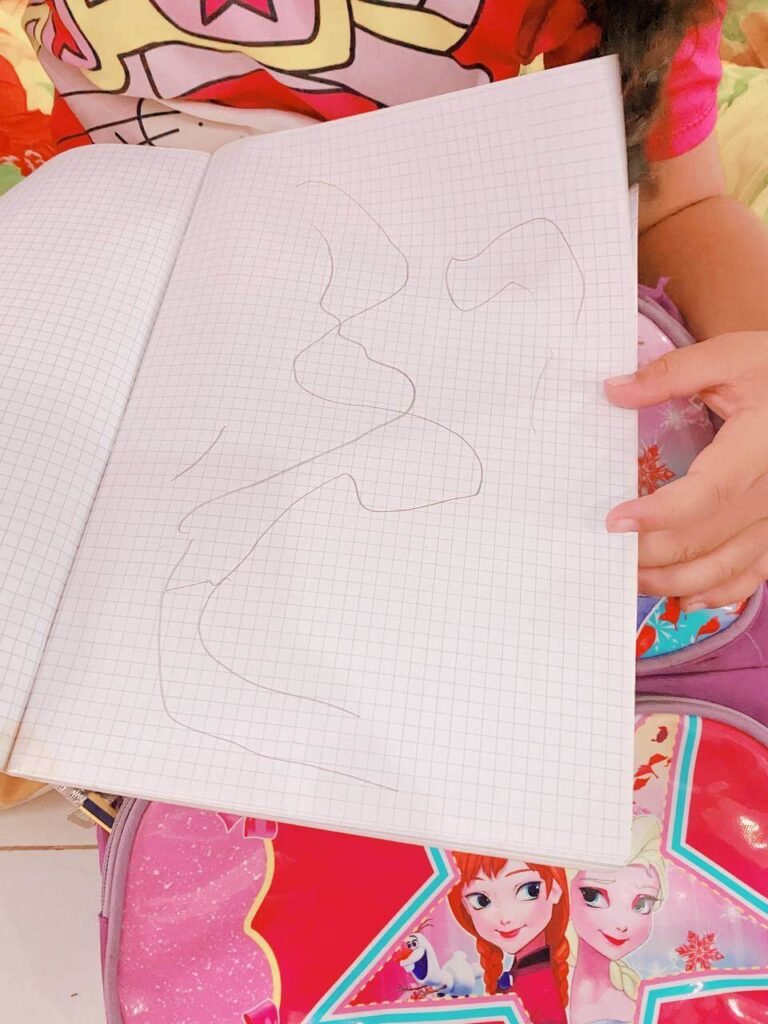 A child's hand holding open a grid notebook showing a hand-drawn, squiggly line map of their indoor obstacle course.
