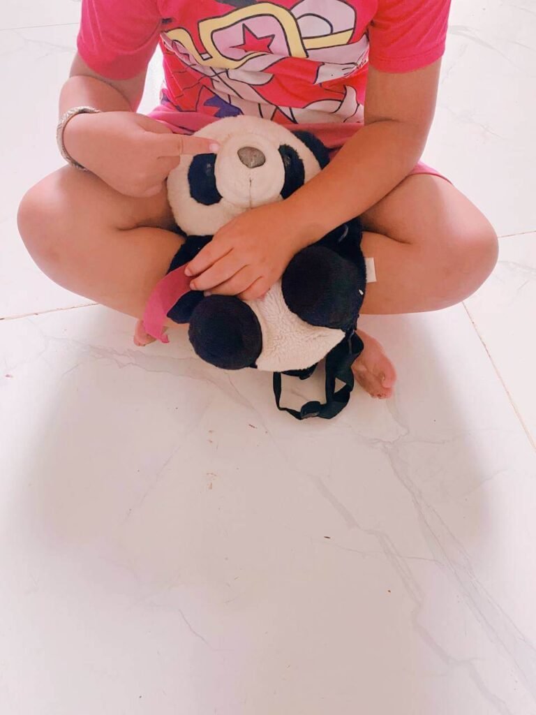 A child sitting cross-legged on the floor, pointing to and hugging a black and white stuffed panda bear toy.