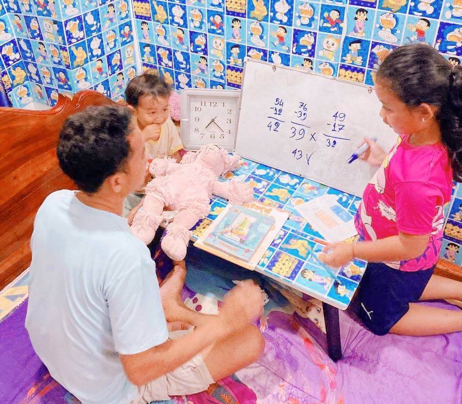 An 8-year-old girl writes math equations on a small whiteboard while her dad and younger brother sit and listen.