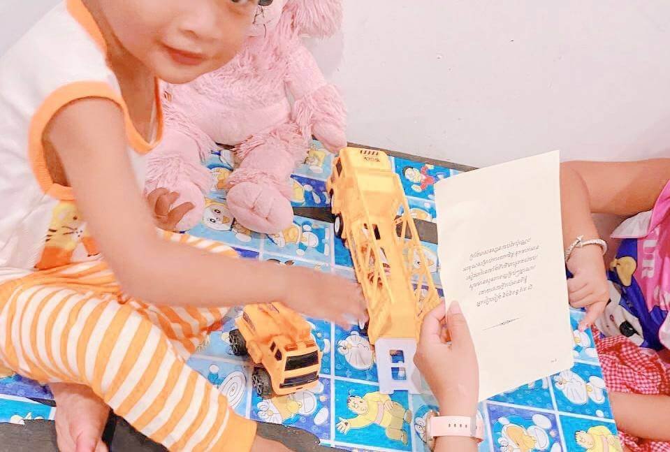 A young boy sitting on a table playing with a yellow toy truck while an adult sitting next to him holds up a piece of paper with text.
