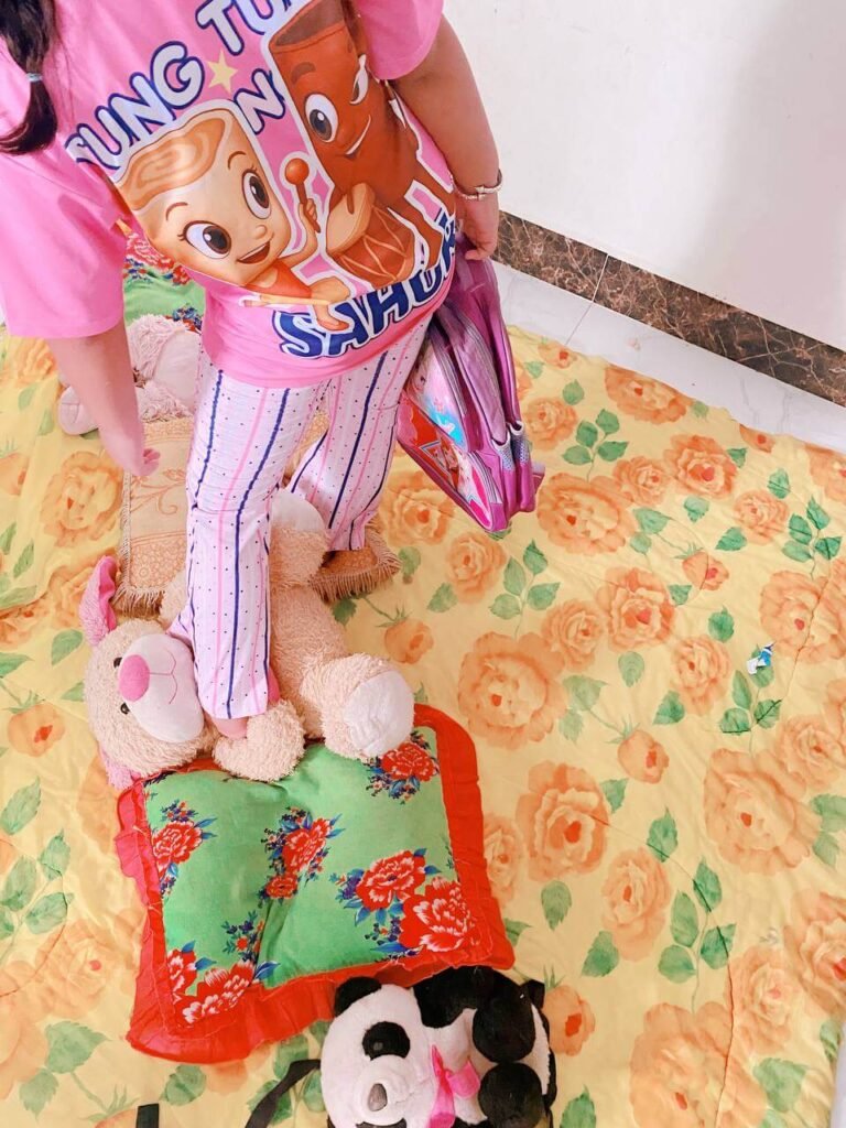 A child stepping onto a large stuffed teddy bear and a floral pillow spread out on a blanket to avoid touching the hard floor.